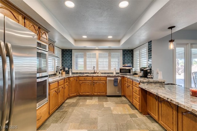 kitchen featuring light stone countertops, stainless steel appliances, brown cabinets, stone finish flooring, and a tray ceiling