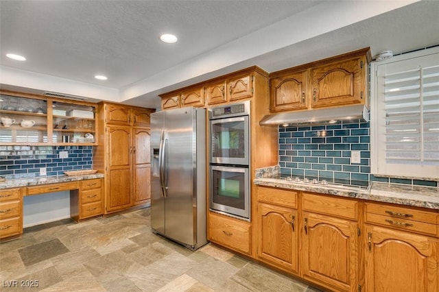 kitchen featuring under cabinet range hood, stainless steel appliances, backsplash, built in study area, and brown cabinets