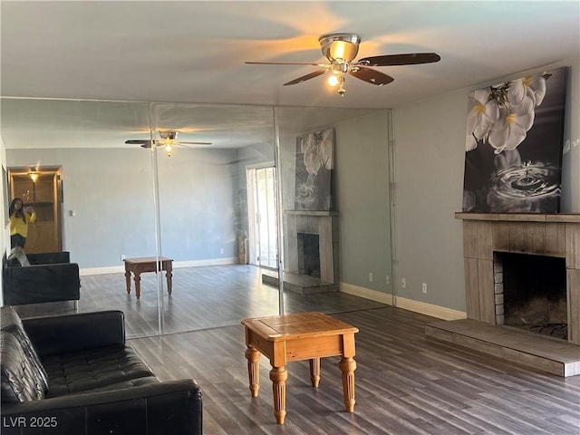 living room with a fireplace with raised hearth, dark wood finished floors, a ceiling fan, and baseboards