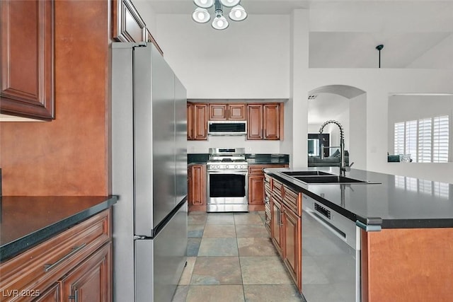 kitchen featuring a center island with sink, brown cabinetry, dark countertops, stainless steel appliances, and a sink