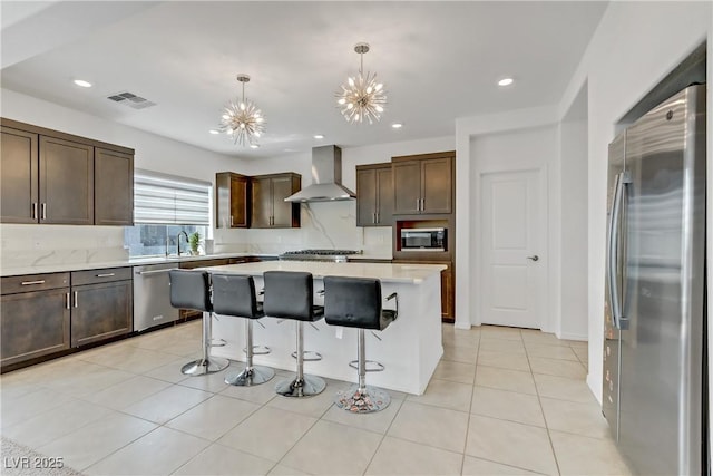 kitchen with stainless steel appliances, dark brown cabinets, wall chimney range hood, hanging light fixtures, and a center island