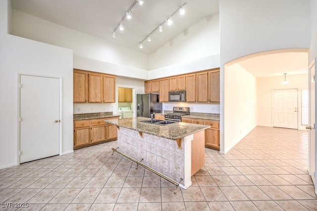 kitchen with arched walkways, light stone counters, a breakfast bar area, black appliances, and a center island with sink