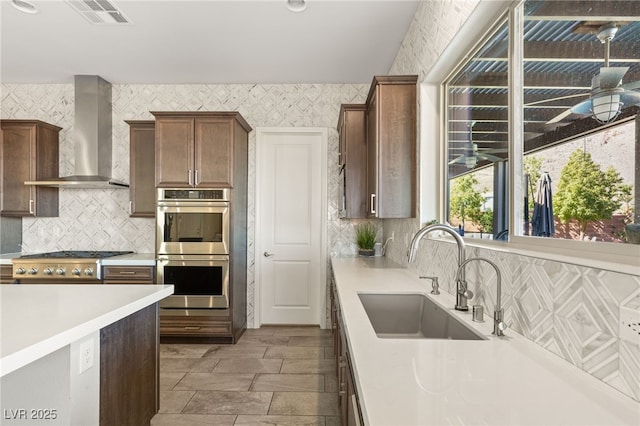 kitchen with stainless steel double oven, a sink, wall chimney range hood, and wallpapered walls