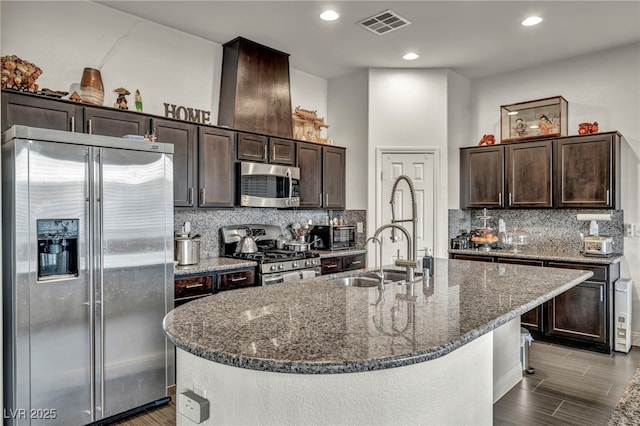 kitchen with an island with sink, dark stone countertops, appliances with stainless steel finishes, and dark brown cabinets