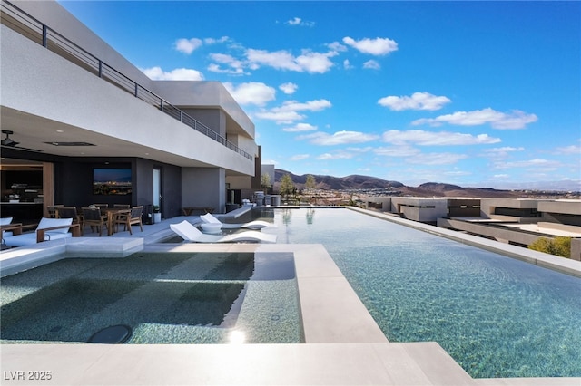 view of swimming pool featuring an infinity pool, a patio, outdoor dining space, ceiling fan, and a mountain view