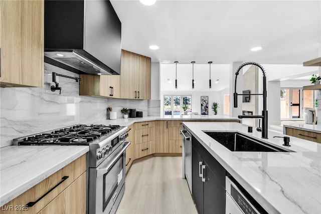 kitchen with light stone counters, stainless steel appliances, hanging light fixtures, a sink, and wall chimney range hood