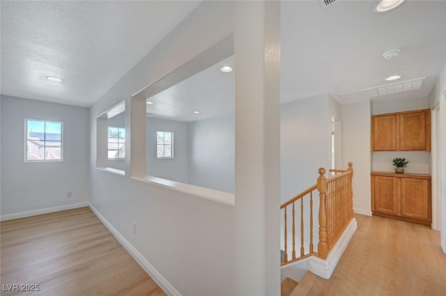 hallway featuring a textured ceiling, visible vents, baseboards, an upstairs landing, and light wood-style floors