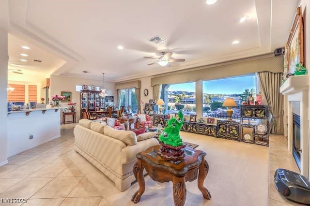 living room featuring light tile patterned floors, ceiling fan with notable chandelier, visible vents, a glass covered fireplace, and crown molding