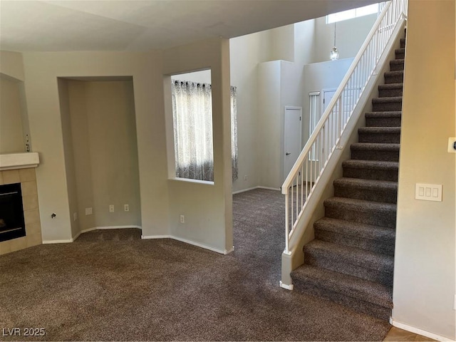 unfurnished living room featuring carpet floors, a wealth of natural light, a tiled fireplace, baseboards, and stairs