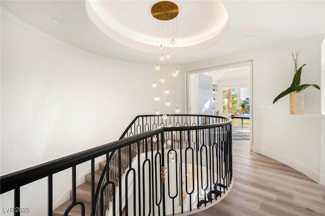 corridor featuring wood finished floors, baseboards, a tray ceiling, recessed lighting, and an upstairs landing