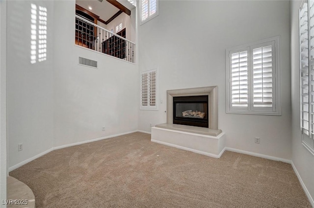 unfurnished living room with a high ceiling, carpet flooring, visible vents, baseboards, and a glass covered fireplace