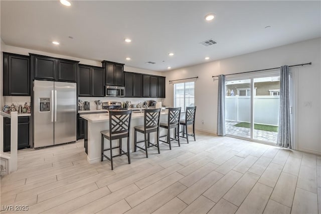 kitchen with visible vents, appliances with stainless steel finishes, a kitchen island with sink, light countertops, and dark cabinetry