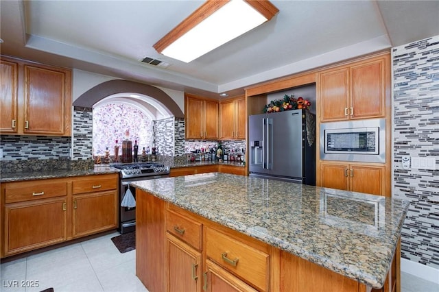 kitchen with light stone counters, appliances with stainless steel finishes, brown cabinetry, a kitchen island, and ventilation hood