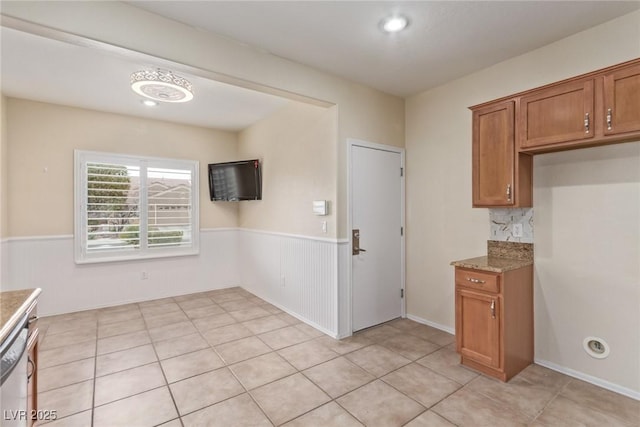 kitchen with a wainscoted wall, brown cabinets, light stone counters, backsplash, and dishwasher