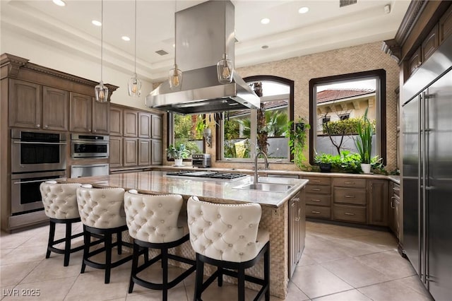 kitchen featuring a kitchen island with sink, island range hood, a sink, appliances with stainless steel finishes, and light stone countertops