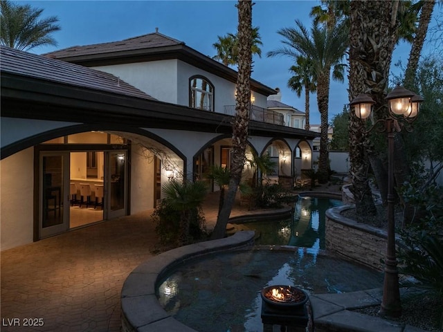 back of property at dusk with a balcony, a patio area, a pool with connected hot tub, and stucco siding