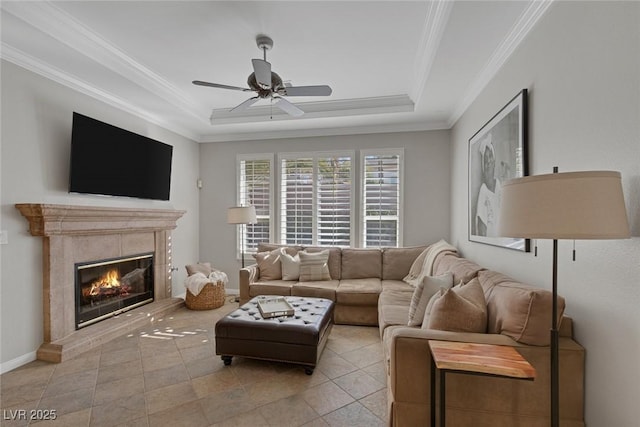 living area featuring a tray ceiling, crown molding, a tiled fireplace, a ceiling fan, and baseboards