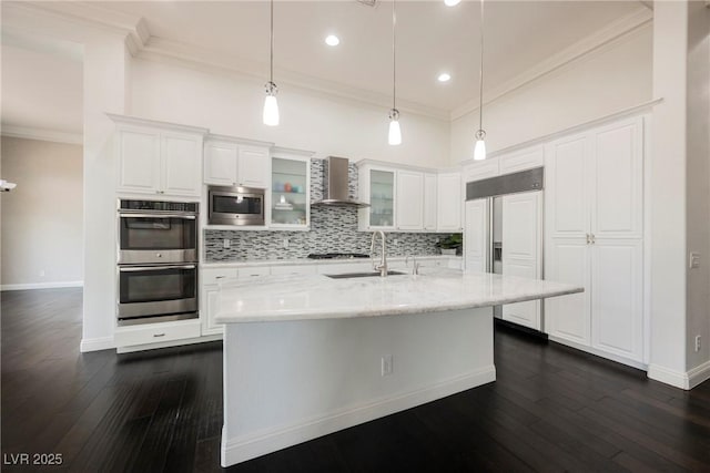 kitchen featuring built in appliances, a sink, white cabinets, wall chimney range hood, and glass insert cabinets