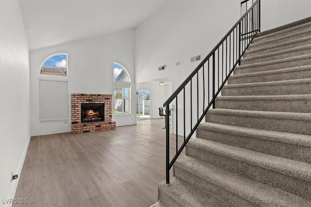 staircase with a brick fireplace, wood finished floors, visible vents, and high vaulted ceiling