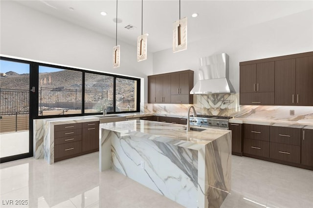 kitchen with decorative light fixtures, a towering ceiling, a kitchen island with sink, a sink, and wall chimney range hood