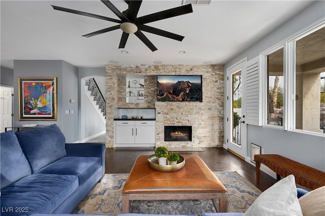 living area featuring dark wood-style flooring, ceiling fan, a stone fireplace, and baseboards