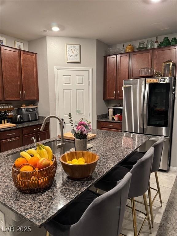 kitchen featuring a breakfast bar area, smart refrigerator, a kitchen island with sink, dark brown cabinetry, and dark stone counters