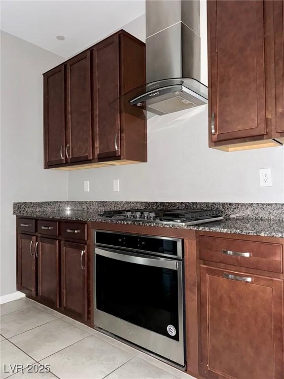kitchen with wall chimney exhaust hood, dark stone countertops, dark brown cabinetry, appliances with stainless steel finishes, and light tile patterned floors