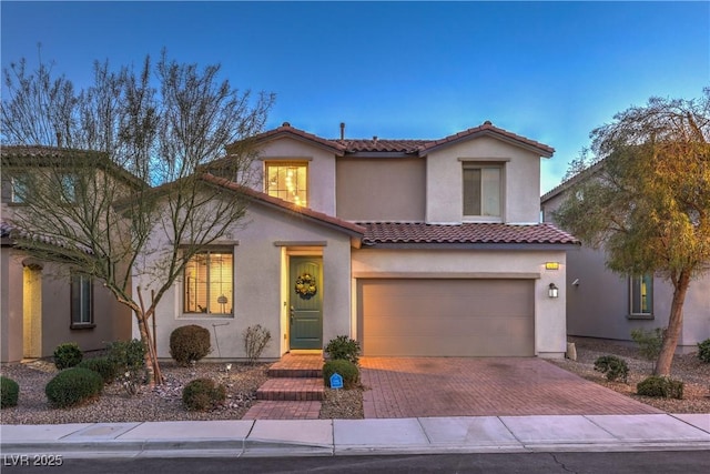 mediterranean / spanish-style house with an attached garage, a tiled roof, decorative driveway, and stucco siding