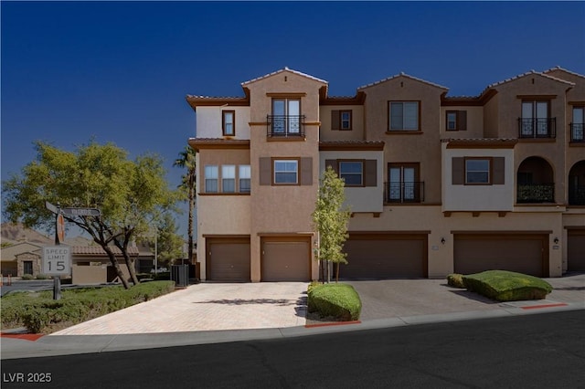 view of front facade featuring a balcony, an attached garage, central AC, stucco siding, and decorative driveway