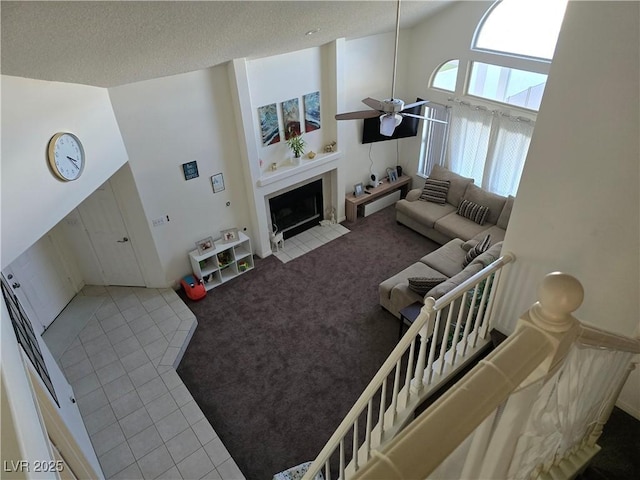 living room with light colored carpet, a high ceiling, a fireplace with flush hearth, a ceiling fan, and a textured ceiling