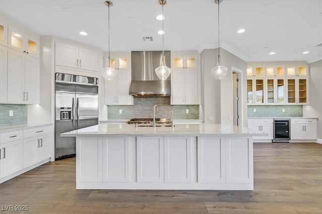 kitchen featuring a kitchen island with sink, beverage cooler, built in refrigerator, wall chimney range hood, and glass insert cabinets