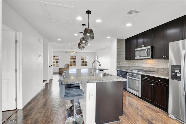 kitchen featuring an island with sink, light stone counters, stainless steel appliances, pendant lighting, and a sink