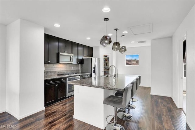 kitchen with stainless steel appliances, a sink, dark stone counters, an island with sink, and decorative light fixtures