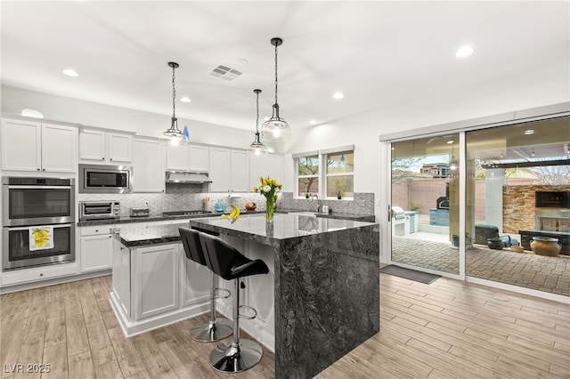 kitchen with under cabinet range hood, a center island, appliances with stainless steel finishes, white cabinets, and decorative backsplash