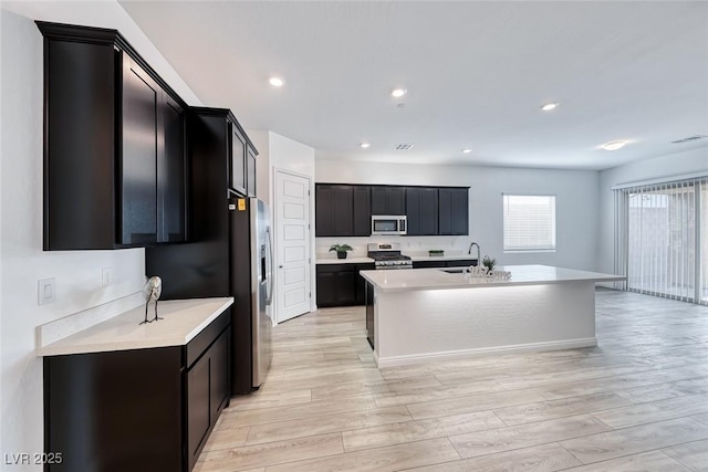 kitchen with appliances with stainless steel finishes, light wood-type flooring, light countertops, and an island with sink