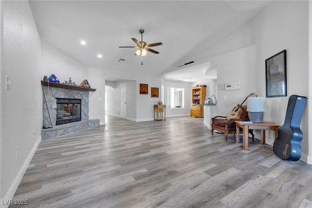 living room with ceiling fan, a stone fireplace, baseboards, vaulted ceiling, and light wood finished floors