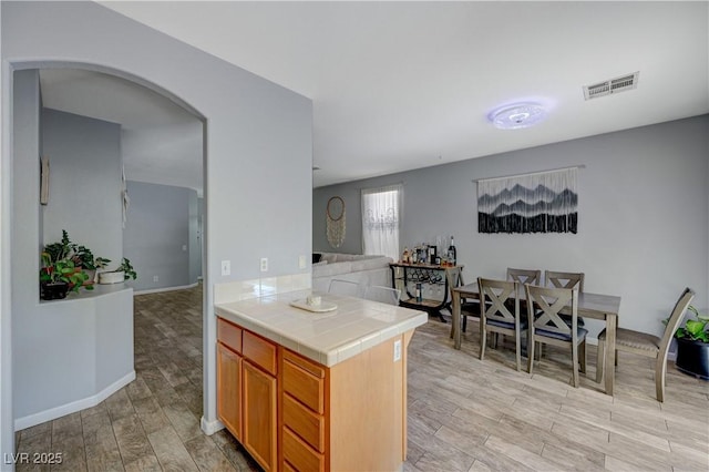 kitchen with tile countertops, arched walkways, light wood-style flooring, visible vents, and brown cabinetry