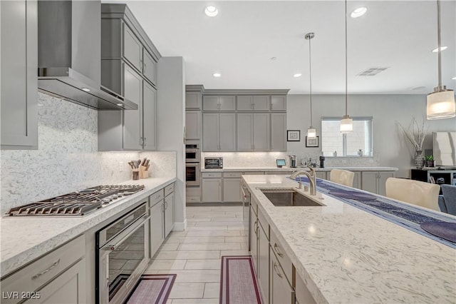 kitchen featuring light stone counters, stainless steel appliances, a sink, gray cabinets, and wall chimney exhaust hood