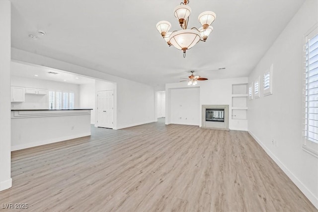 unfurnished living room featuring built in features, light wood finished floors, a glass covered fireplace, baseboards, and ceiling fan with notable chandelier