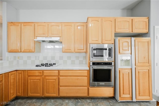 kitchen featuring tile countertops, backsplash, light brown cabinetry, appliances with stainless steel finishes, and under cabinet range hood