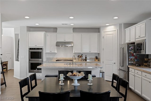 kitchen featuring light stone counters, a center island with sink, appliances with stainless steel finishes, white cabinetry, and under cabinet range hood