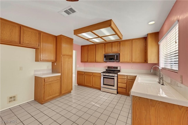 kitchen with a sink, visible vents, appliances with stainless steel finishes, tile counters, and brown cabinets