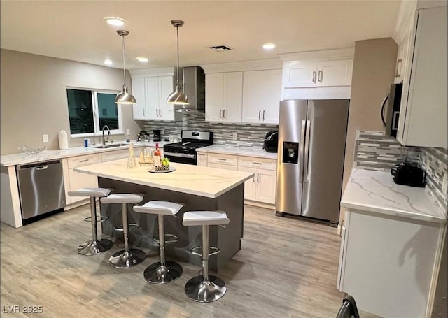 kitchen featuring hanging light fixtures, appliances with stainless steel finishes, a sink, wall chimney range hood, and a kitchen island