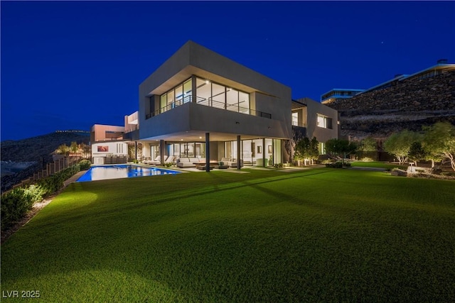 back of house at night featuring a balcony, a patio area, an outdoor pool, and a yard