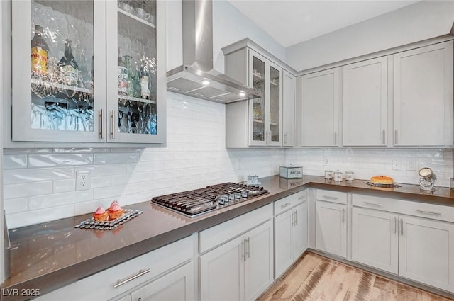 kitchen with decorative backsplash, dark countertops, glass insert cabinets, wall chimney range hood, and stainless steel gas cooktop