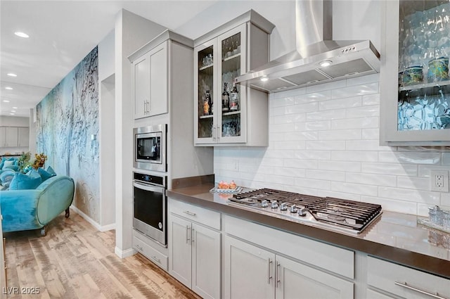 kitchen with appliances with stainless steel finishes, glass insert cabinets, light wood-style floors, and wall chimney range hood