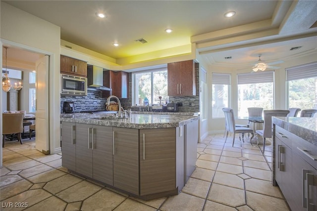 kitchen with tasteful backsplash, brown cabinetry, wall chimney exhaust hood, stainless steel microwave, and a sink