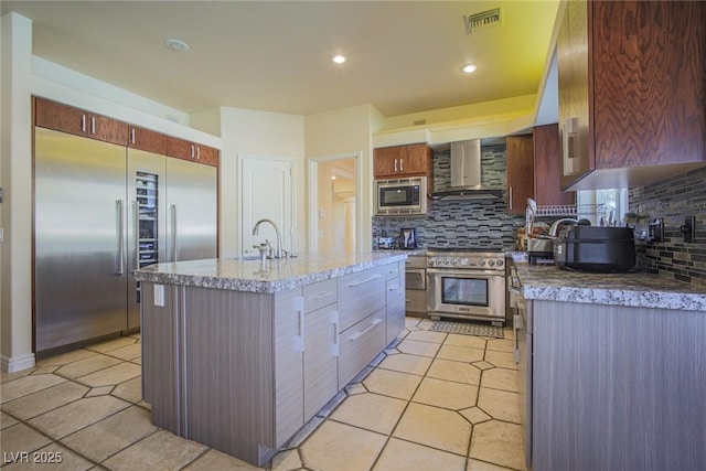 kitchen featuring built in appliances, a kitchen island with sink, a sink, decorative backsplash, and wall chimney exhaust hood