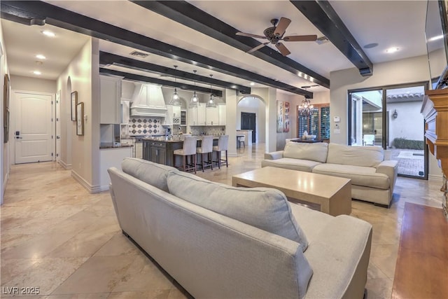 living room featuring arched walkways, visible vents, beamed ceiling, baseboards, and ceiling fan with notable chandelier