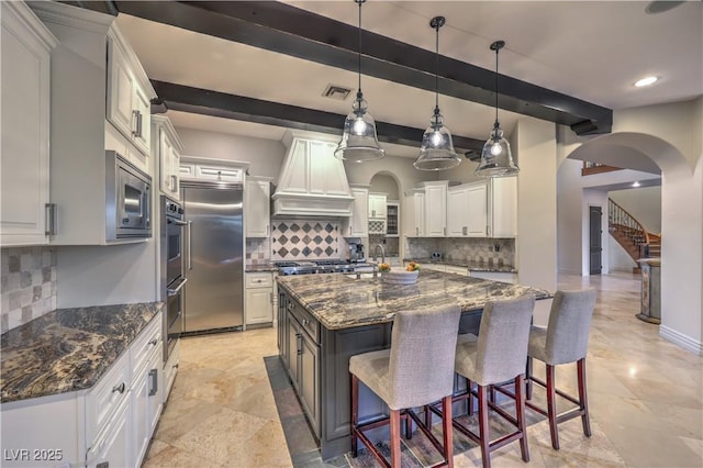 kitchen featuring visible vents, an island with sink, custom range hood, a kitchen breakfast bar, and built in appliances